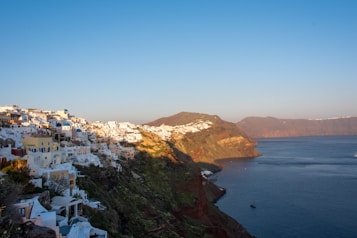 A picturesque view of whitewashed buildings with blue domes perched on a cliff overlooking the calm blue sea. The warm glow of the setting sun illuminates the landscape, highlighting the rugged terrain and the contrast between the bright buildings and the natural surroundings.