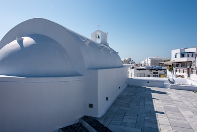 A whitewashed building with a domed roof, typical of Cycladic architecture, sits under a clear blue sky. A small cross adorns the peak of the structure, and nearby, a sign indicates the presence of a supermarket. The area is paved with light-colored stones, and other similar structures are visible in the background.