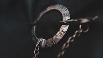 A close-up of a textured metal brooch with intricate details, featuring a circle and a pin. The craftsmanship highlights a hammered surface, with a chain hanging from it, set against a dark background.