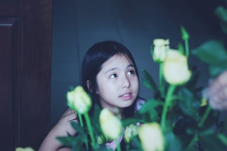 A young girl with long dark hair is looking upwards, surrounded by several yellow roses with green leaves. The background is dimly lit, highlighting her face and the flowers in the foreground.