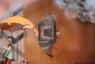 A metal wind spinner with geometric, square shapes hangs in the foreground, creating a sense of depth and motion. In the background, there is a blurred, whimsical figure of a person holding an umbrella with bright colors.
