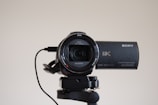 A high-definition Sony 4K Handycam mounted on a tripod, with the lens facing forward. The camcorder features a Zeiss lens and is connected by a cable. It appears clean and ready for use, with details of its specifications visible on the camera body.