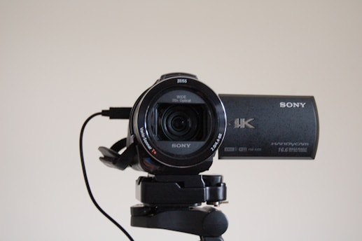 A high-definition Sony 4K Handycam mounted on a tripod, with the lens facing forward. The camcorder features a Zeiss lens and is connected by a cable. It appears clean and ready for use, with details of its specifications visible on the camera body.