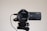 A high-definition Sony 4K Handycam mounted on a tripod, with the lens facing forward. The camcorder features a Zeiss lens and is connected by a cable. It appears clean and ready for use, with details of its specifications visible on the camera body.