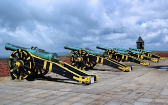 A row of historical cannons mounted on ornate wheeled carriages is positioned on a brick-paved platform. The cannons are distinguished by their turquoise barrels and black carriages with yellow accents. The background features a partly cloudy sky, and a small structure with a conical roof is visible to the right.