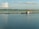 A tranquil body of water with a small floating fish farm consisting of blue barrels and wooden structures. A few people are in a boat near one of the structures, and the horizon is lined with trees under a sky dotted with clouds.