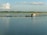 A tranquil body of water with a small floating fish farm consisting of blue barrels and wooden structures. A few people are in a boat near one of the structures, and the horizon is lined with trees under a sky dotted with clouds.