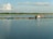 A tranquil body of water with a small floating fish farm consisting of blue barrels and wooden structures. A few people are in a boat near one of the structures, and the horizon is lined with trees under a sky dotted with clouds.