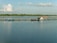 A tranquil body of water with a small floating fish farm consisting of blue barrels and wooden structures. A few people are in a boat near one of the structures, and the horizon is lined with trees under a sky dotted with clouds.