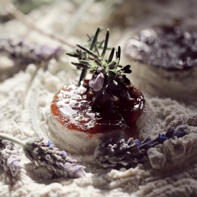A soft cheese topped with a rich red jelly and garnished with sprigs of herbal greenery and purple flowers. The cheese rests on a powdery white surface, surrounded by additional purple flowers and herbs, creating a rustic and artisanal presentation.