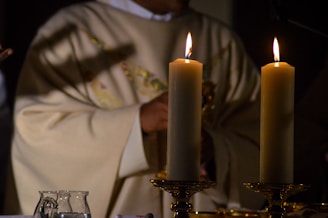 Two lit candles are placed on ornate holders with a person wearing a ceremonial robe in the background, creating a warm and solemn atmosphere. The robe has embroidered patterns and a subtle shadow of the cross is visible.