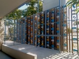 A modern metal gate in front of an artistic installation made of vertical wooden panels with mounted colorful cylindrical tubes. The tubes are blue, white, and yellow, arranged in a pattern. The structure is surrounded by greenery and sunlight casts shadows on the platform below.