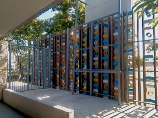 A modern metal gate in front of an artistic installation made of vertical wooden panels with mounted colorful cylindrical tubes. The tubes are blue, white, and yellow, arranged in a pattern. The structure is surrounded by greenery and sunlight casts shadows on the platform below.