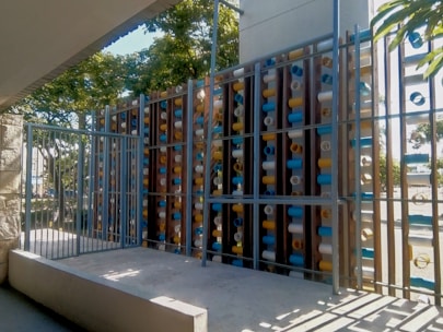 A modern metal gate in front of an artistic installation made of vertical wooden panels with mounted colorful cylindrical tubes. The tubes are blue, white, and yellow, arranged in a pattern. The structure is surrounded by greenery and sunlight casts shadows on the platform below.