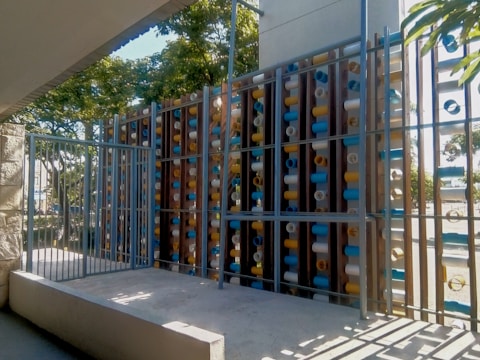 A modern metal gate in front of an artistic installation made of vertical wooden panels with mounted colorful cylindrical tubes. The tubes are blue, white, and yellow, arranged in a pattern. The structure is surrounded by greenery and sunlight casts shadows on the platform below.