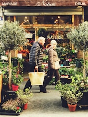 An outdoor flower shop with vibrant plants and flowers arranged in pots, framed by two trees. Two people are walking in front, wearing winter clothing and carrying bags. The shop's awning displays the words 'Nature' and related terms.