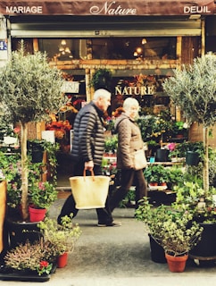 An outdoor flower shop with vibrant plants and flowers arranged in pots, framed by two trees. Two people are walking in front, wearing winter clothing and carrying bags. The shop's awning displays the words 'Nature' and related terms.
