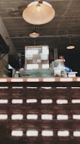 A rustic interior setting featuring vintage lighting fixtures hanging from the ceiling. A retro-style cash register sits on a counter, with scattered items including a large glass jar. The backdrop consists of a textured brick wall with light patterns creating a unique visual effect.