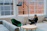 A modern, minimalist interior features a woman sitting on a white chair, using a laptop. The room includes a gray sofa, a green chair, and a wooden coffee table. Large windows with white frames line the walls, revealing a view of red and blue brick exteriors. A black floor lamp arches over the seating area, set in a space with white walls and radiators.