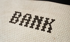 The word 'BANK' is spelled out in uppercase letters using dark hexagonal tiles against a white hexagonal tile floor.