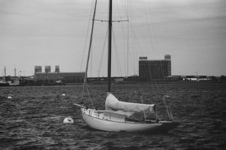 A sailboat with a covered sail is moored on a body of water. In the background, there are several large buildings and a ship labeled 'NANTUCKET'. The sky is overcast, contributing to a monochromatic and subdued atmosphere.