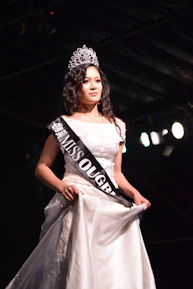 Close-up of a crowned winner smiling confidently, holding a bouquet on stage with a sparkling backdrop.