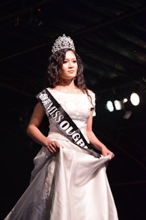 A woman wearing a white gown and a crown stands on a stage. She has a sash across her chest with 'MISS OUGRI 2018' printed on it. The background is dark with stage lighting highlighting her.