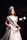 A woman wearing a white gown and a crown stands on a stage. She has a sash across her chest with 'MISS OUGRI 2018' printed on it. The background is dark with stage lighting highlighting her.