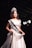 A woman wearing a white gown and a crown stands on a stage. She has a sash across her chest with 'MISS OUGRI 2018' printed on it. The background is dark with stage lighting highlighting her.