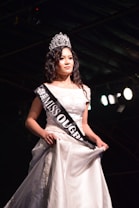 A woman wearing a white gown and a crown stands on a stage. She has a sash across her chest with 'MISS OUGRI 2018' printed on it. The background is dark with stage lighting highlighting her.