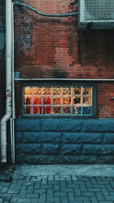 A brick wall with a rectangular glass block window showing warm lights shining through, possibly suggesting an interior space with vibrant elements. The lower part of the wall is painted in a dark blue tone, while a section of the wall features an air conditioning unit and several pipes.