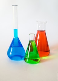 Three laboratory glassware items are filled with colorful liquids. On the left, a long-necked blue flask, in the middle a small green beaker, and on the right a red conical flask. They are placed on a plain white surface against a soft, neutral background.