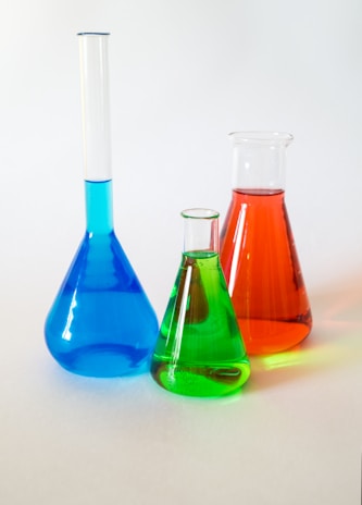 Three laboratory glassware items are filled with colorful liquids. On the left, a long-necked blue flask, in the middle a small green beaker, and on the right a red conical flask. They are placed on a plain white surface against a soft, neutral background.