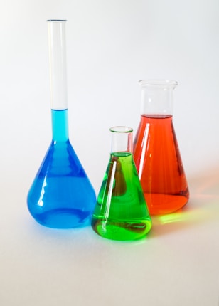 Three laboratory glassware items are filled with colorful liquids. On the left, a long-necked blue flask, in the middle a small green beaker, and on the right a red conical flask. They are placed on a plain white surface against a soft, neutral background.