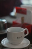 A ceramic cup sits on a saucer with steam rising, indicating a hot beverage inside. There's a logo on the cup, and the background features blurred objects with hints of red, suggesting a cozy indoor setting.