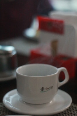 A ceramic cup sits on a saucer with steam rising, indicating a hot beverage inside. There's a logo on the cup, and the background features blurred objects with hints of red, suggesting a cozy indoor setting.