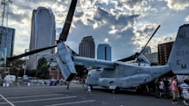 A military aircraft is parked in a cityscape setting with tall buildings forming the background. Several people, including adults and children, are gathered around the aircraft. The sky above features dramatic clouds with rays of sunlight peeking through, creating a striking contrast.