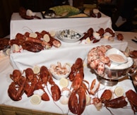 A lavish seafood display features multiple lobsters, large shrimp, a whole fish, lemon slices, and bowls of sauce. The arrangement is on a white tablecloth, suggesting a formal dining or buffet setting.