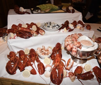 A lavish seafood display features multiple lobsters, large shrimp, a whole fish, lemon slices, and bowls of sauce. The arrangement is on a white tablecloth, suggesting a formal dining or buffet setting.