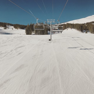 A snowy ski slope stretches out under a clear blue sky, with ski lifts overhead and evergreen trees lining the sides. The snow has visible ski tracks and is well-groomed.