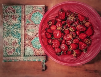 A woven textile with intricate geometric patterns in teal and brown lies next to a red plastic basket filled with ripe strawberries. The strawberries appear fresh and vibrant, with visible seeds and green leaves, contrasting against the ornate design of the fabric.