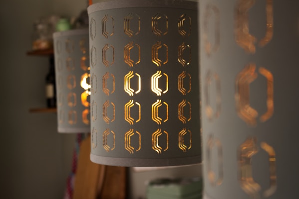 A series of cylindrical lampshades with geometric cutouts that reveal warm, glowing light. The details of the cutouts are intricate, and the light creates an inviting and cozy atmosphere. The background is softly blurred, with shelves containing objects like bottles and containers.