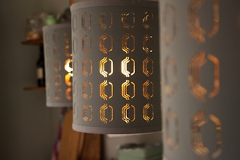 A series of cylindrical lampshades with geometric cutouts that reveal warm, glowing light. The details of the cutouts are intricate, and the light creates an inviting and cozy atmosphere. The background is softly blurred, with shelves containing objects like bottles and containers.