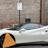 A white sports car is parked on the street with a wheel clamp on its front left tire. The background features a brick wall with a sign on a lamp post, indicating parking restrictions.