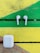 Two white wireless earbuds are placed on a wooden surface painted with vibrant green and yellow colors. Below them is a white charging case.