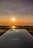 A sleek iPhone is placed on a surface with a stunning sunset in the background. The sun glows brightly, casting warm hues across the sky and reflecting on a body of water below.