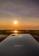 A sleek iPhone is placed on a surface with a stunning sunset in the background. The sun glows brightly, casting warm hues across the sky and reflecting on a body of water below.