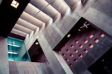 An architectural interior featuring modern design elements with concrete walls and a series of small, square windows that allow light to enter. A glass partition with a door is visible on the left side, and fluorescent lights can be seen reflecting on the surfaces. The interplay of light and shadow creates a dramatic appearance.