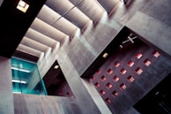 An architectural interior featuring modern design elements with concrete walls and a series of small, square windows that allow light to enter. A glass partition with a door is visible on the left side, and fluorescent lights can be seen reflecting on the surfaces. The interplay of light and shadow creates a dramatic appearance.