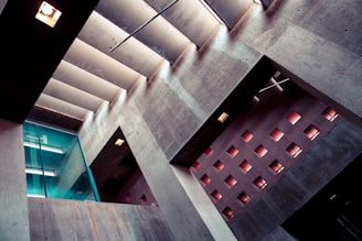 An architectural interior featuring modern design elements with concrete walls and a series of small, square windows that allow light to enter. A glass partition with a door is visible on the left side, and fluorescent lights can be seen reflecting on the surfaces. The interplay of light and shadow creates a dramatic appearance.
